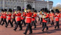 “Marching band of the Coldstream Guards in red tunics performing in front of Buckingham Palace during the Changing of the Guard ceremony.”